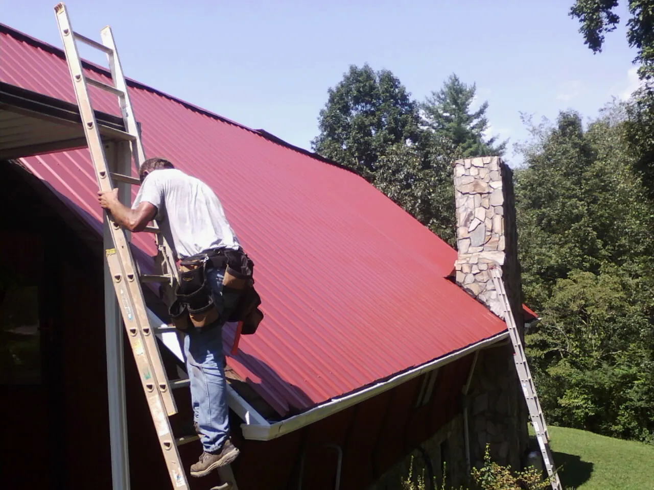 Roofer on a ladder beside a red metal roof and stone chimney — Soffit & Fascia Repair by Roofing Brothers in Schoharie